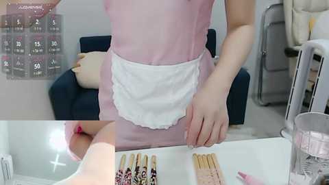 Media: Video of an Asian woman in a pink maid outfit with white apron, standing at a white counter. Background shows a dark blue chair, a calendar, and a glass of water.