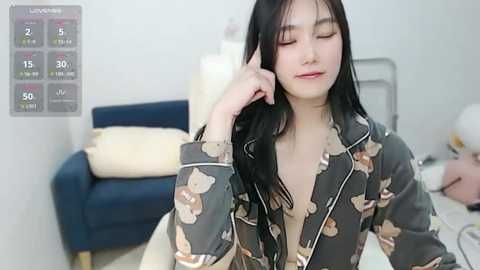 Media: A video of an Asian woman with long black hair, wearing a dark pajama set with teddy bear prints, smiling and talking on her phone, in a modern bedroom with a blue couch, white walls, and a calendar on the left.