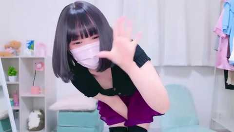 Media: Video of a young woman with a straight bob haircut, wearing a black dress, purple skirt, and a face mask, making a peace sign in a softly lit, pastel-themed room.