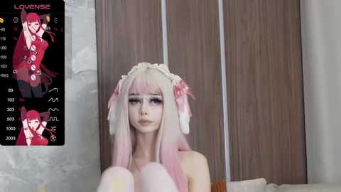 Media: Video of a young woman with long, pastel pink hair in a maid outfit, wearing a headband, sitting on a bed with wooden paneling background.