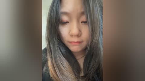 Media: Video of an Asian woman with long, straight, dark brown hair, light skin, wearing a black top, looking down, set against a blurred, neutral-toned background.