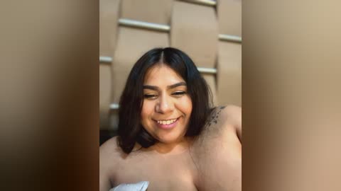 Media: Video of a Latina woman with medium skin tone, shoulder-length black hair, and a tattoo on her right shoulder, smiling while topless, against a beige padded wall.