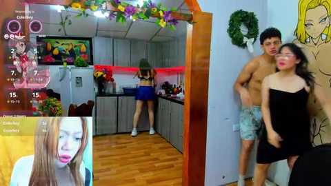 Media: Video: A young woman in a black dress stands beside two shirtless men in a colorful, playful kitchen. A TV screen in the foreground shows a close-up of another woman.