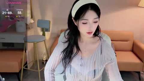 Media: Video of a young East Asian woman with long black hair, wearing a white headband and blouse, sitting in a modern, beige-lit living room with a TV screen displaying a video feed.