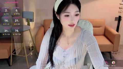 Media: Video of a young Asian woman with long black hair, wearing a light-colored blouse, seated in a modern living room with beige furniture and a green lamp.