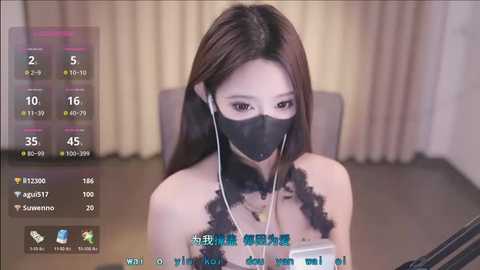 Media: A video of an East Asian woman with long black hair and a black face mask, wearing a white T-shirt, sitting at a desk with a computer. The background features beige curtains.