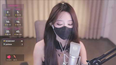 Media: Video of an Asian woman with long, straight black hair singing into a microphone while wearing a black face mask and a black top. Background includes a gray chair and beige curtains.