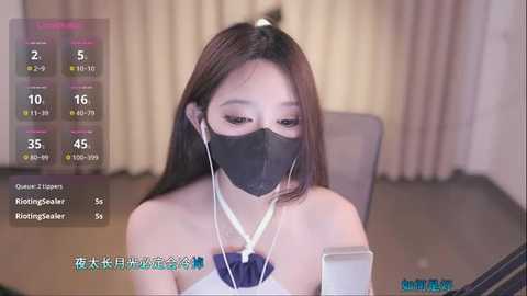 Media: Video of an East Asian woman with long brown hair, wearing a black face mask, seated at a desk, using a smartphone. Background shows curtains and a table.