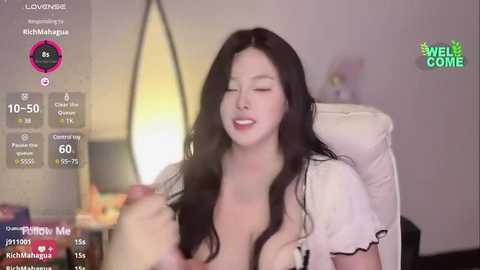 Media: A video of a smiling East Asian woman with long black hair, wearing a white top, holding a phone, in a cozy room.