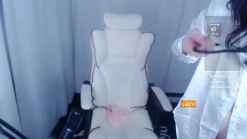 Media: Video of a person with a light-skinned penis and testicles in a white recliner chair, taken in a dimly lit room with a medical theme.