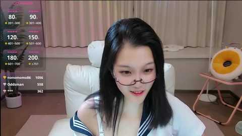 Media: A video of an Asian woman with black hair, wearing glasses, a sailor collar top, and sitting on a white couch in a modern room with beige curtains and a pink chair.