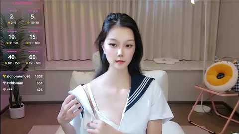 Media: A video of an East Asian woman with long black hair, wearing a sailor uniform, sitting on a white couch in a modern room with a window and a plant.