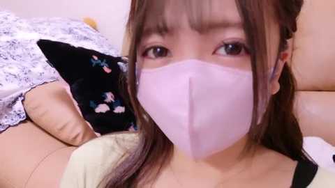 Media: A video of an Asian woman with light skin, brown hair, and bangs, wearing a light pink face mask, lying on a bed with white and floral-patterned sheets, looking directly at the camera.