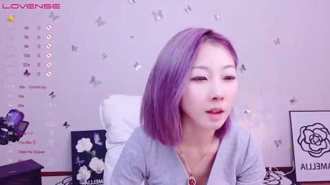 Media: Video of a young Asian woman with light skin and purple hair, smiling, wearing a gray cardigan, in a room with a white wall adorned with butterfly decals and floral artwork, and a camera on a tripod.