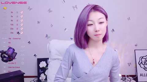 Media: Video of a young Asian woman with shoulder-length purple hair, wearing a light gray V-neck top, sitting in a white chair against a pastel purple wall adorned with butterflies and floral decals.