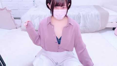 Media: Video of an Asian woman with short black hair, wearing a pink button-up shirt, white pants, and a face mask, kneeling on a white floor in a minimalist bedroom.