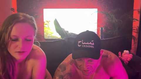 Media: Video of a darkly lit room with a man in a black cap and a woman with long hair, both shirtless, watching a TV screen.