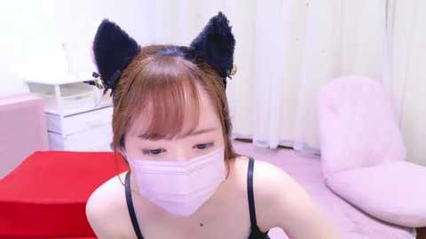 Media: Video of a young Asian woman in a cat-themed outfit, including black cat ears and a face mask, sitting on a red cushion. Background includes white curtains and a pink chair.