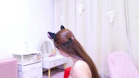 Media: Video of a woman with long brown hair, wearing black cat ears, facing away, in a white room with a vanity table and a pink chair.