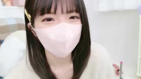 Media: Video of an East Asian woman with straight, dark brown hair and bangs, wearing a pink face mask and white top, seated indoors with blurred background.