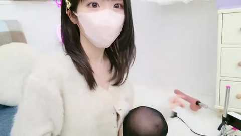 Media: Video of an Asian woman with straight black hair, wearing a white mask, sitting in a minimalist room with white walls, holding a microphone, and using a hairdryer.