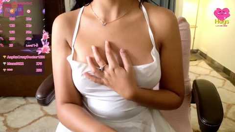 Media: Video of a woman with medium skin tone, wearing a white tank top, sitting in a chair, covering her breasts with her hands, in a modern room with a floral-patterned wall.