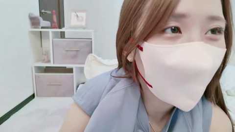 Media: Video of an Asian woman with light skin and brown hair in a medical mask, wearing a light blue shirt, in a clean, white room with storage shelves and pastel-colored bins.