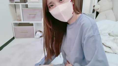 Media: Video of an Asian woman with long brown hair, wearing a white face mask, blue sweatshirt, and sitting on a white bed in a tidy, white-walled bedroom with pink storage drawers and plush toys.