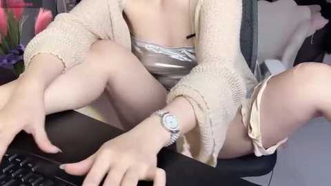 Media: Video of a woman in a beige crochet cardigan, a silver bra, and light shorts, typing on a black keyboard, legs crossed, with a watch on her wrist.