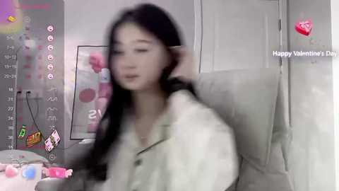 Media: A blurred photo of an Asian woman with long black hair, wearing a white top, standing in a room with pink decorations.