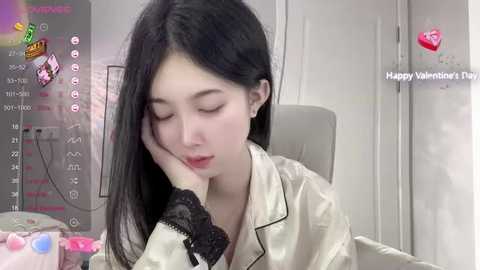 Media: A video of an Asian woman with long black hair, wearing a silky beige robe, resting her head on her hand, indoors with a Valentine's Day message in the background.