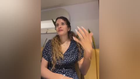 Media: Video of a curvy woman with long, wavy hair, wearing a navy blue dress with white polka dots, gesturing with her right hand. She stands in a modern, beige-toned room with white walls and a large mirror.