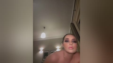 Media: Video of a topless woman with fair skin, blonde hair, and heavy makeup, blowing a kiss. She is in a dimly lit room with a white ceiling and framed artwork in the background.