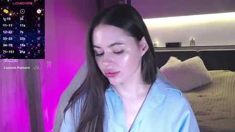 Media: Video of a young woman with long black hair, wearing a light blue shirt, standing in a modern bedroom with a bed, purple lighting, and a nightstand.