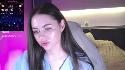 Media: Video of a fair-skinned woman with long brown hair, wearing a light blue sweater, in a cozy bedroom with a dark sofa, a nightstand, and a purple and pink LED light.