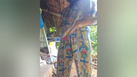 Media: A video of a person in bright blue and yellow Doraemon pajama pants, bending over under a thatched roof, surrounded by greenery and a rustic outdoor setting.