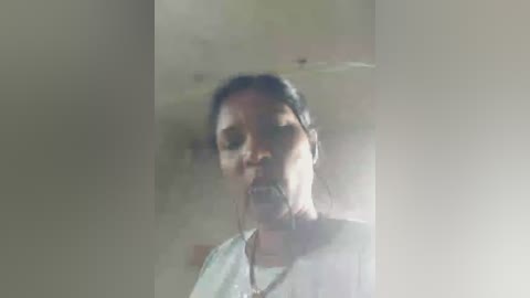 Media: A blurry video of a middle-aged woman with dark skin and short hair, wearing a white shirt, standing in a dimly lit room with a window and a light source creating a halo effect around her head.