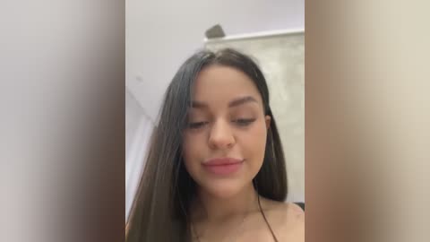 Media: A video of a young woman with long, straight black hair, light skin, and full lips, wearing a black top, looking downward with a serene expression. The background is blurred, featuring a white wall and a glimpse of a gray surface.