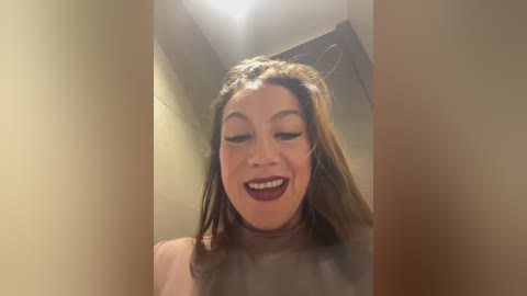 Media: Video of a smiling woman with light skin, brown hair, and dark red lipstick, wearing a light-colored top, captured from a slightly tilted angle.