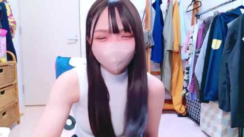 Media: Video of a young Asian woman with long black hair, wearing a white sleeveless top, with a blurred face, taking a selfie in a cluttered, brightly lit bedroom with clothes hanging on racks.