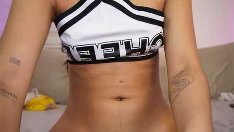 Media: Video of a light-skinned woman wearing a white and black \"Cheetahs\" sports bra, revealing toned abs and tattoos on her arms. Background shows a beige couch and blurred room details.