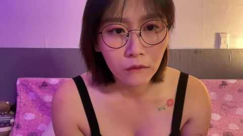 Media: Video of a young East Asian woman with short black hair, wearing round glasses, black tank top, and floral pillowcase, seated indoors.