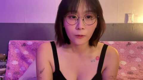 Media: Video of a young Asian woman with short black hair, wearing glasses, black tank top, and floral-patterned pillow. She has tattoos on her shoulders and is seated on a bed with a pink and white floral bedspread.