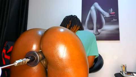 Media: Video of a nude Black woman with dark skin and long braids, bent over, receiving a butt plug, in a room with a black-and-white painting of a woman sitting on a bed.