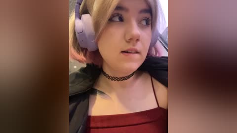 Media: Video of a young woman with short blonde hair, wearing a black choker and red tank top, sitting in a car with large pink headphones, blurred background.