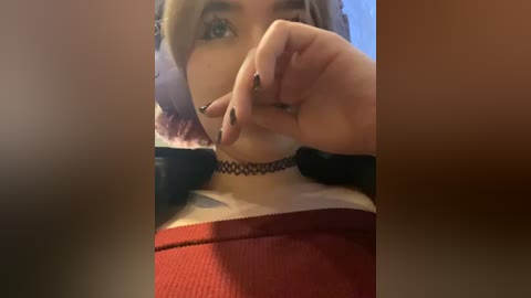 Media: Video of a young woman with light skin and short, wavy blonde hair, wearing a black choker necklace, a red sweater, and black nail polish. She's partially covering her face with her hand.