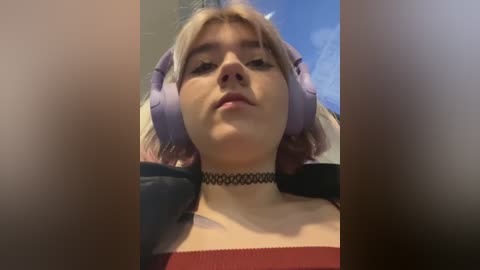 Media: A video of a young woman with light skin, blonde hair, and a black choker, wearing a red top and large purple headphones. The background shows a cloudy sky.