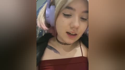 Media: Video of a young woman with pale skin, platinum blonde hair, and large, purple headphones, wearing a black choker and a maroon tank top, smiling while eyes are closed.