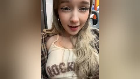 Media: A video of a young woman with long, wavy blonde hair, wearing a white tank top with the word \"DICK\" and a plaid shirt, seated in a car with blurred background.