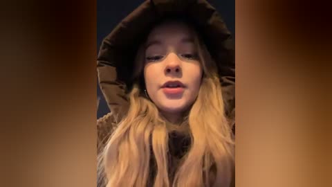 Media: Video of a young woman with long, blonde hair, wearing a brown puffy coat, with a serene expression against a blurred, dark background.